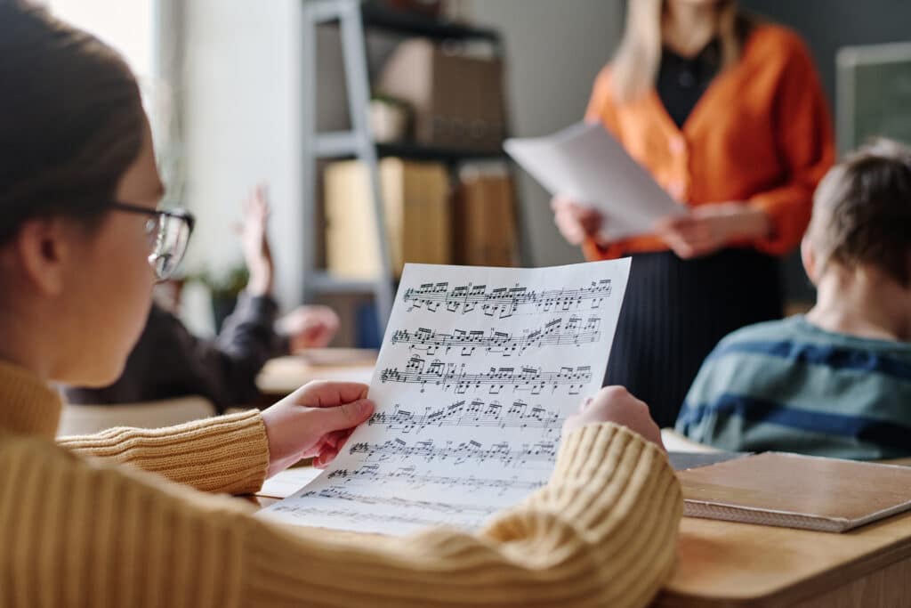 Unrecognizable girl wearing eyeglasses sitting at desk in classroom holding sheet music paper and listening to teacher during music lesson