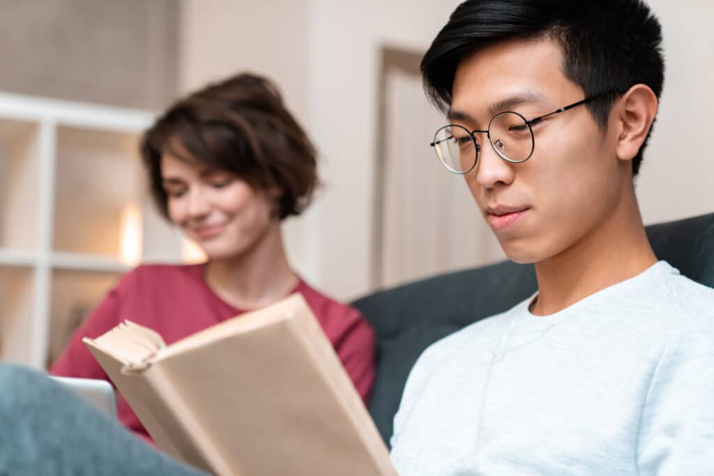 Photo of pleased multinational couple reading book and using laptop while sitting on sofa at home